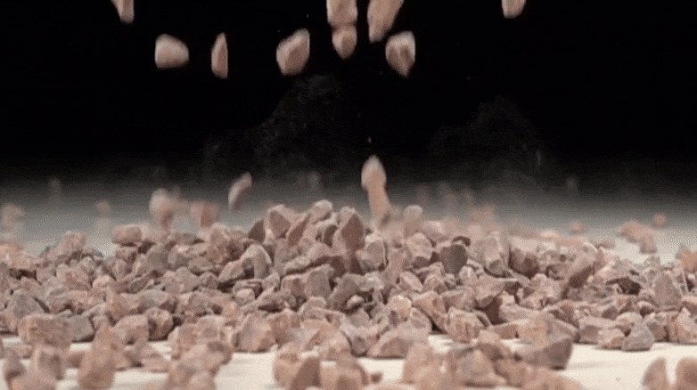 Mehrere kleine, rötliche Steine fallen aus der Luft herab und bilden einen Haufen auf einer hellen Oberfläche. Der Hintergrund ist dunkel, sodass die Bewegung der Steine und der entstehende Steinhaufen im Vordergrund deutlich hervorgehoben werden.

                          